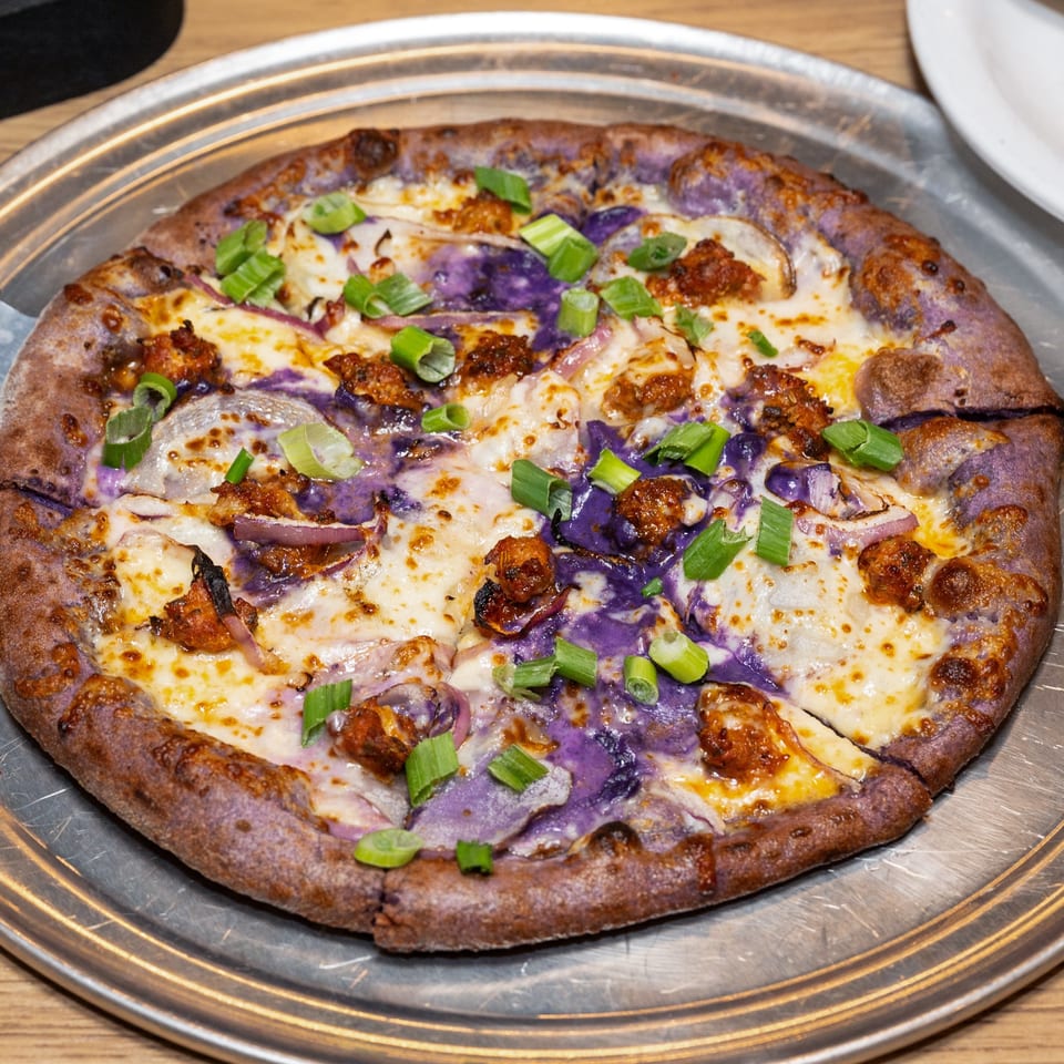 Purple-crusted pizza with melted cheese, purple sauce, caramelized onions, and green onions on a metal tray.