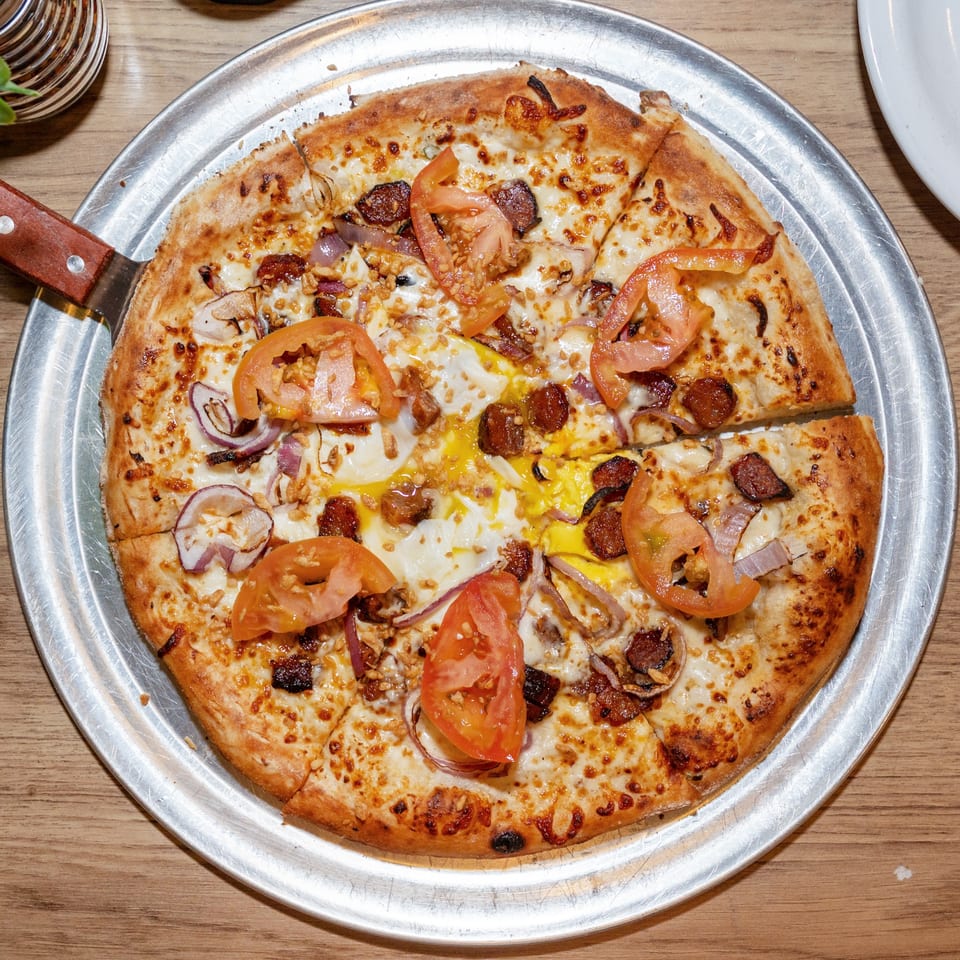 A sliced pizza with tomatoes, onions, bacon, and a fried egg on a metal tray.