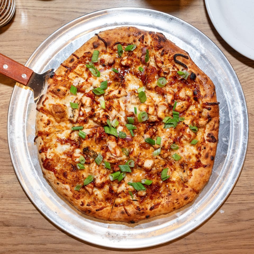 A BBQ chicken pizza with melted cheese and green onions on a metal tray with a pizza server.