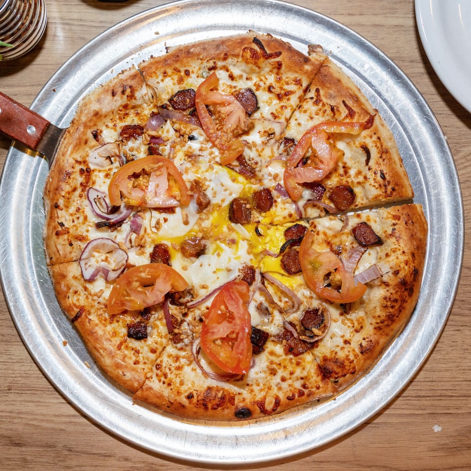 A freshly baked pizza with tomatoes, onions, sausage, and eggs on a metal tray.