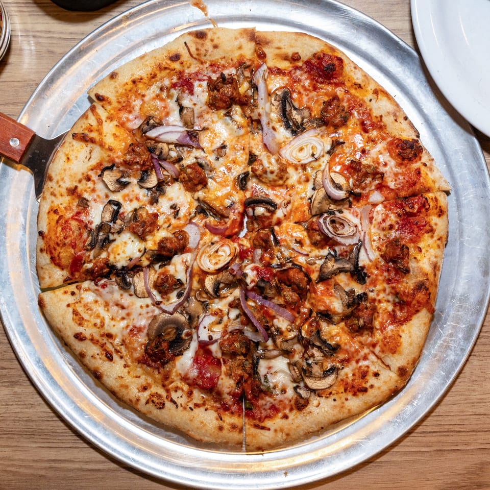 A whole pizza with sausage, mushrooms, and red onions on a metal tray with a server.
